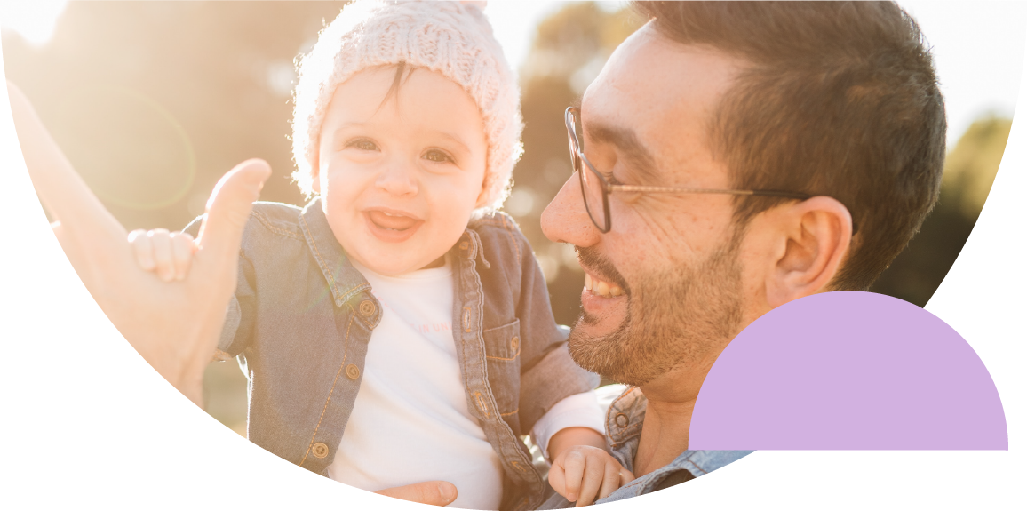 Un papá alza a su beba, ambos sonriendo, en un campo iluminado por el sol de la tarde.